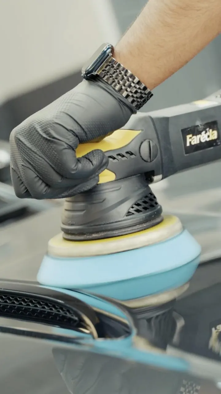 A gloved hand guiding a dual-action polisher with a blue pad across a car&rsquo;s paintwork